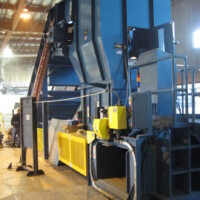 Industrial recycling machine with blue and yellow panels inside a large warehouse, featuring a ladder and control panels.