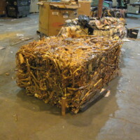 A large, compacted bale of mixed scrap metal, including copper, is placed on a warehouse floor.
