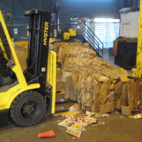 Forklift moving a large bale of compressed cardboard in a recycling facility, with loose papers scattered on the floor.