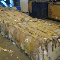 Large compacted block of flattened cardboard boxes in a recycling facility, surrounded by loose cardboard pieces.