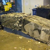 Compacted bundle of crushed metal and cardboard near a baling machine inside an industrial facility.