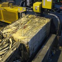 A machine compresses a stack of recycled paper and cardboard bound with metal ties in an industrial setting.