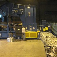A worker operates a large blue industrial machine in a factory, with processed metal pieces exiting on a conveyor belt.