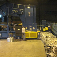 A worker operates a large blue industrial machine in a factory, with processed metal pieces exiting on a conveyor belt.