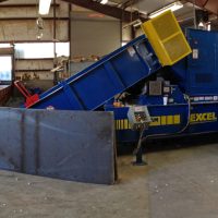 Industrial paper baler machine in a warehouse, surrounded by stacks of compressed paper bales and loose materials.