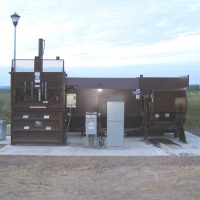Large brown metal trash compactor and recycling unit on a concrete platform outdoors, with a lamp post and grass in the background.